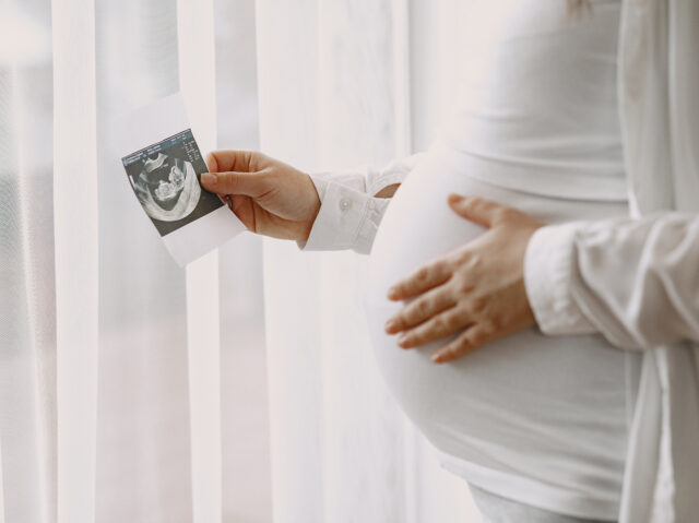 Pregnant woman stands by the window and looks at photo